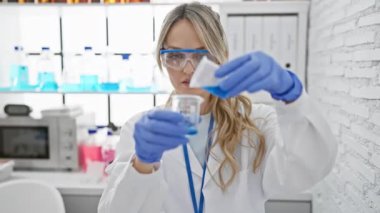 Laboratuvar önlüklü beyaz kadın bilim adamı modern bir laboratuvarda mavi bir sıvıyla deney yapıyor.