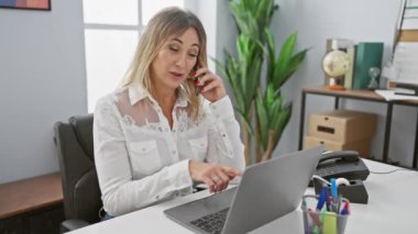 Profesyonel bir kadın telefonda, modern bir ofiste dizüstü bilgisayarı, masası ve ofis malzemeleriyle konuşuyor..