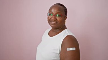 Kendinden emin Afro-Amerikalı kadın, neşeli bir şekilde gülümsüyor, kolundaki yara bandına işaret ediyor.