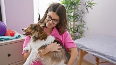 Pembe önlüklü gülümseyen genç bir kadın veteriner kliniğinde üç renkli bir köpek tutuyor..