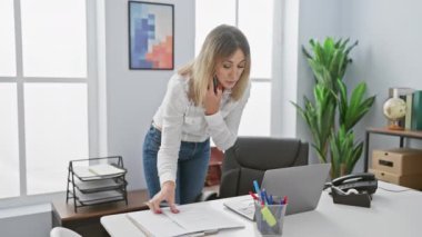 Modern bir ofiste birden fazla iş yapan olgun sarışın bir kadın. Elinde dizüstü bilgisayar ve belgelerle telefonda konuşurken masaya yaslanıyor..