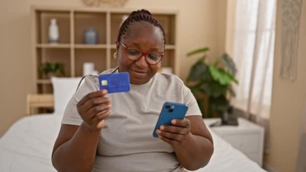 彼女の寝室に快適に座っているアフリカ系アメリカ人女性の笑顔 彼女の居心地の良い屋内部屋でクレジットカードを使用して スマートフォンで朝のオンラインショッピングを楽しむ — ストック動画