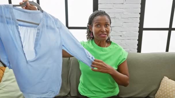 成熟したアフリカの女性は 明るいリビングルームで思慮深い表情のソファに座って屋内で青いシャツを検査します — ストック動画
