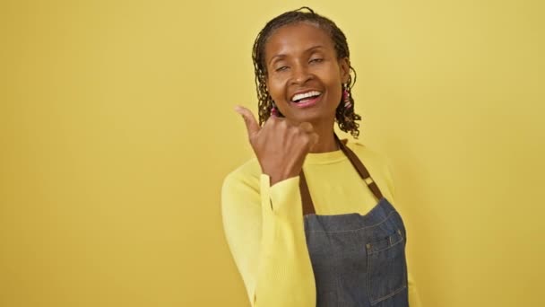 エプロンの陽気な中年アフリカ系アメリカ人女性は 活気に満ちた黄色い背景に孤立した親指で 喜んで側を指しています — ストック動画