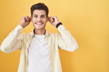 Sarı arka planda dikilen İspanyol genç adam gülümsüyor parmaklarıyla kulaklarını çekiyor, komik bir jest. deneme sorunu 