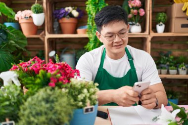 Young chinese man florist smiling confident using smartphone at flower shop