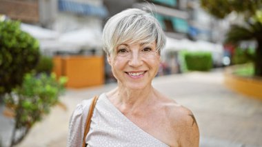 Güneşli bir caddede, açık havada gülümseyen gri kısa saçlı, olgun İspanyol bir kadın.