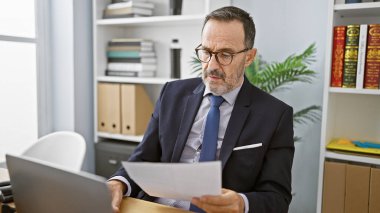 Olgun ve odaklanmış orta yaşlı iş adamı, gri saçlı, ofisinde bir iş belgesi okuyor, işine odaklanmış.
