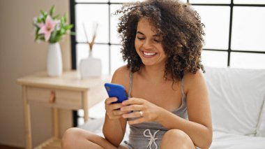 Genç, güzel, İspanyol bir kadın yatakta akıllı telefon kullanıyor.