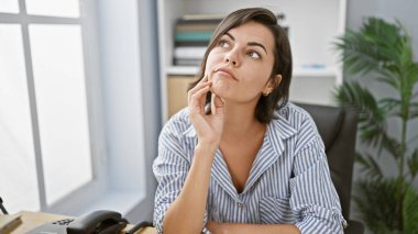 İş yerinde, genç, güzel İspanyol kadın çalışan, ciddi düşüncelerle büyülenmiş, ofis masasında oturan, iş başarısına odaklanmış bir portre..