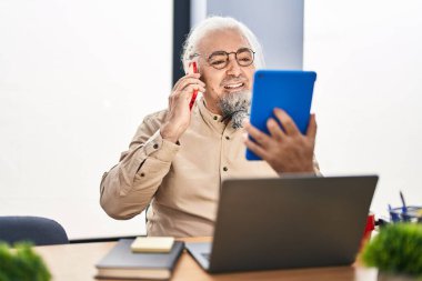 Middle age grey-haired man business worker talking on smartphone using touchpad at office