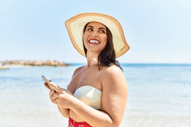 Genç, güzel İspanyol kadın turist deniz kenarında akıllı telefon kullanarak gülümsüyor.