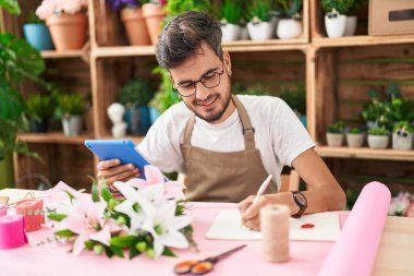 Young hispanic man florist using touchpad writing on notebook at flower shop