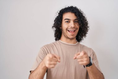 Beyaz arka planda duran kıvırcık saçlı İspanyol adam seni ve kamerayı işaret ediyor, gülümsüyor, pozitif ve neşeli. 