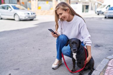 Akıllı telefon kullanan genç sarışın kadın sokak köpeğinin yanında duruyor.