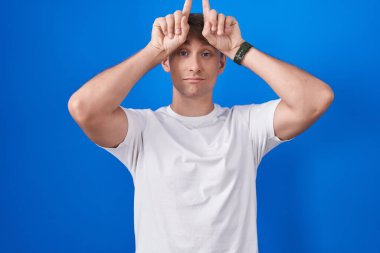 Kafkasyalı sarışın adam mavi arka planda duruyor ve boynuz gibi parmağıyla komik jestler yapıyor. 