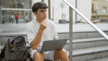 Düşüncelere dalmış, genç, yakışıklı İspanyol bir öğrenci, dizüstü bilgisayarına dalmış, şehir üniversitesinin merdivenlerinde rahatça oturuyor.