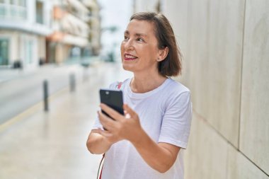 Sokakta akıllı telefon kullanan orta yaşlı bir kadın gülümsüyor.