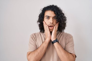 Beyaz arka planda duran kıvırcık saçlı İspanyol adam korkmuş ve şok olmuş, şaşırmış ve şaşırmış bir yüz ifadesi var. 