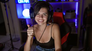 Alıngan genç İspanyol kadın flaması, güzel kısa saçlı, oyununa devam ediyor! Oyun odasında video oynuyorum, başparmak hareketi yapıyorum, konsolunun ışıltısıyla gülümsüyorum.