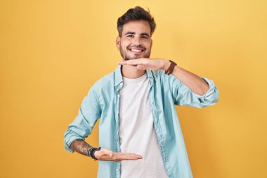 Young hispanic man with tattoos standing over yellow background gesturing with hands showing big and large size sign, measure symbol. smiling looking at the camera. measuring concept. 