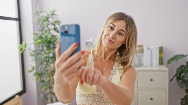 Sarışın kadın evinde selfie çekiyor. Parlak bir iç mekanda yaşam tarzı, teknoloji ve zarafet sergiliyor..