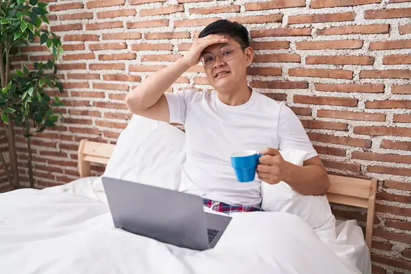 Young asian man drinking coffee sitting on the bed stressed and frustrated with hand on head, surprised and angry face