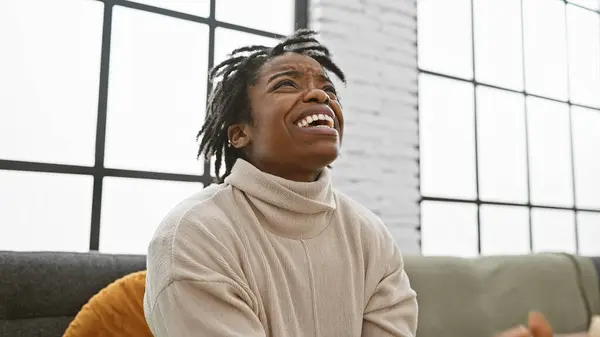 Portrait of a laughing young black woman with dreadlocks at home on a sofa, showcasing emotion and indoor lifestyle.