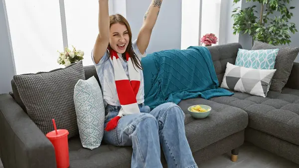 Excited woman cheering on sofa at home, surrounded by snacks and cozy cushions.
