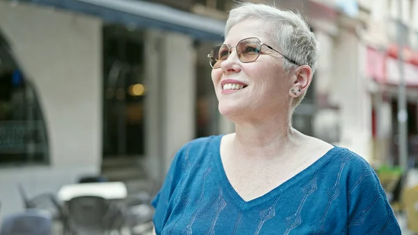 Middle age grey-haired woman wearing sunglasses looking to the sky smiling at street