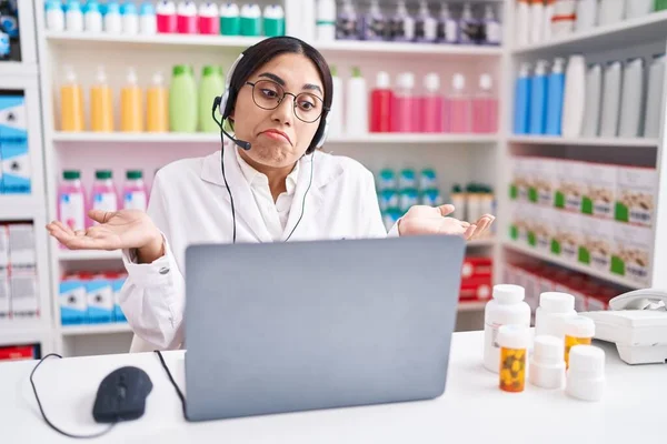 Young arab woman working at pharmacy drugstore using laptop clueless and confused expression with arms and hands raised. doubt concept.
