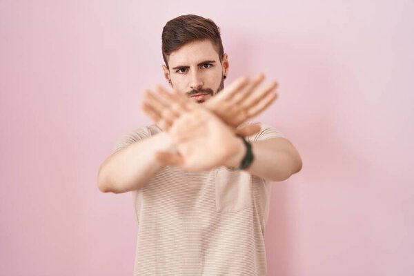 Hispanic man with beard standing over pink background rejection expression crossing arms and palms doing negative sign, angry face 
