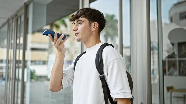 Confident young hispanic teenager, a smart university student, casually standing and effortlessly sending an audio message via smartphone amidst busy campus life, his backpack slung over one shoulder.