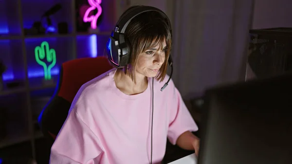 Beautiful young hispanic woman streamer, seriously immersed in a cyber game, sits relaxed at night playing on her computer in the gaming room, live streaming with short hair, headphones & microphone