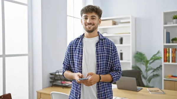 Confident young arab man, beaming with a happy expression, engrossed in work, effortlessly manages business tasks on his smartphone, standing in an elegant office indoors.