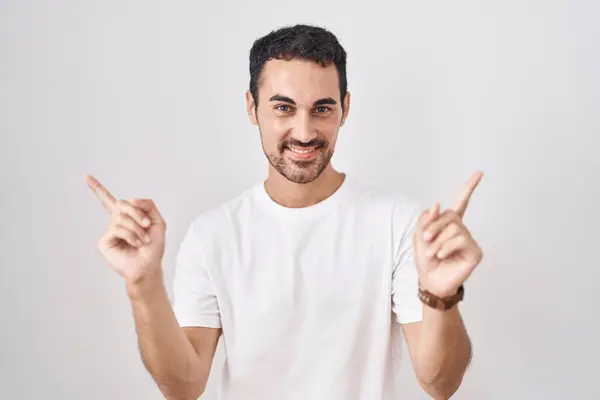 白い背景の上に立っているハンサムなヒスパニックの男性は 異なる方向に指で指を指して自信を持って微笑みます 広告のためのコピースペース — ストック写真