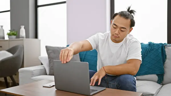 Handsome young chinese man, relaxed yet concentrated, opening his laptop while sitting on the sofa at home. indoor shot of asian adult using technology to work online in his living room.
