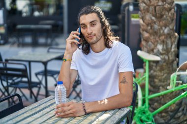 Young hispanic man talking on smartphone sitting on table at coffee shop terrace