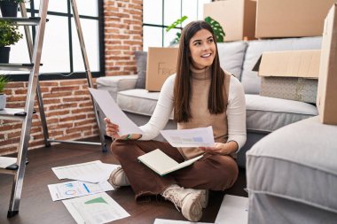 Young beautiful hispanic woman reading document sitting on floor at new home