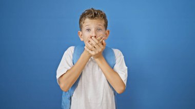 Sevimli sarışın öğrenci şaşkınlık içinde, ağzı kapalı, mavi arka planda çalışma çantasıyla ayakta duruyor.