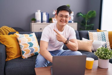 Genç Asyalı adam evinde dizüstü bilgisayar kullanıyor. Kanepede mutlu bir şekilde oturuyor. Yüzünde mutlu ve doğal bir ifadeyle elleri ve parmaklarıyla işaret eden bir gülümsemeyle. 