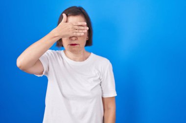 Middle age hispanic woman standing over blue background covering eyes with hand, looking serious and sad. sightless, hiding and rejection concept 
