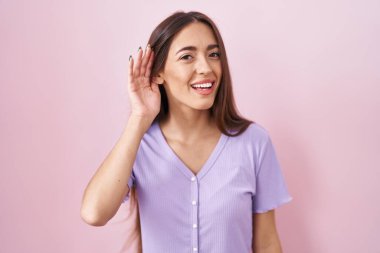 Pembe arka planda duran uzun saçlı, ağzı kulaklarında dedikoduları ve dedikoduları dinleyen genç İspanyol kadın. Sağırlık kavramı. 