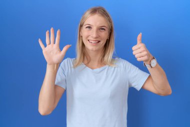 Kafkasyalı genç bir kadın günlük mavi tişört giyiyor. Altıncı parmaklarını gösterip gösterirken kendinden emin ve mutlu gülümsüyor.. 