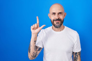 Mavi arka planda dikilen İspanyol dövmeli adam iki numarayı gösterip işaret ederken kendinden emin ve mutlu bir şekilde gülümsüyor.. 