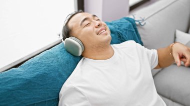 Mutlu Çinli adam, evde müzik dinlerken neşeyle dans ediyor. Oturma odasındaki kanepede kulaklıklarıyla oturuyor.