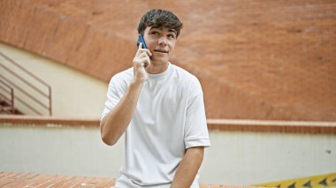 Gündüz Latin kökenli genç bir erkek, güneşli bir şehir sokağında ciddi bir telefon konuşmasına dalmış, havalı bir ifadeyle genç bir adamın hayat tarzı portresi, açık havada akıllı telefonuyla tutkuyla konuşuyor.