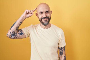 Sarı arka planda dikilen İspanyol dövmeli adam gülümseyerek tek parmağıyla kafasını işaret ediyor, harika bir fikir ya da düşünce, iyi bir hafıza 