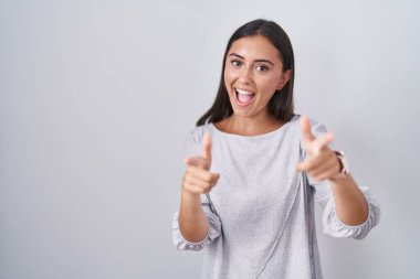 Beyaz arka planda duran genç İspanyol kadın mutlu ve komik yüzlü parmaklarını kameraya doğrultuyor. iyi enerji ve titreşimler. 