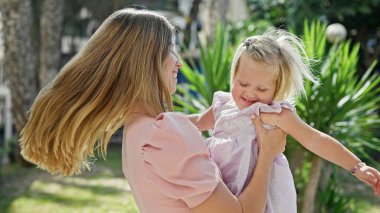 Kendine güveni tam bir anne, kollarını kızına dolamış, neşeyle gülüyor, güneşli parkta duruyor, pozitif enerji ve mutluluk saçıyor.